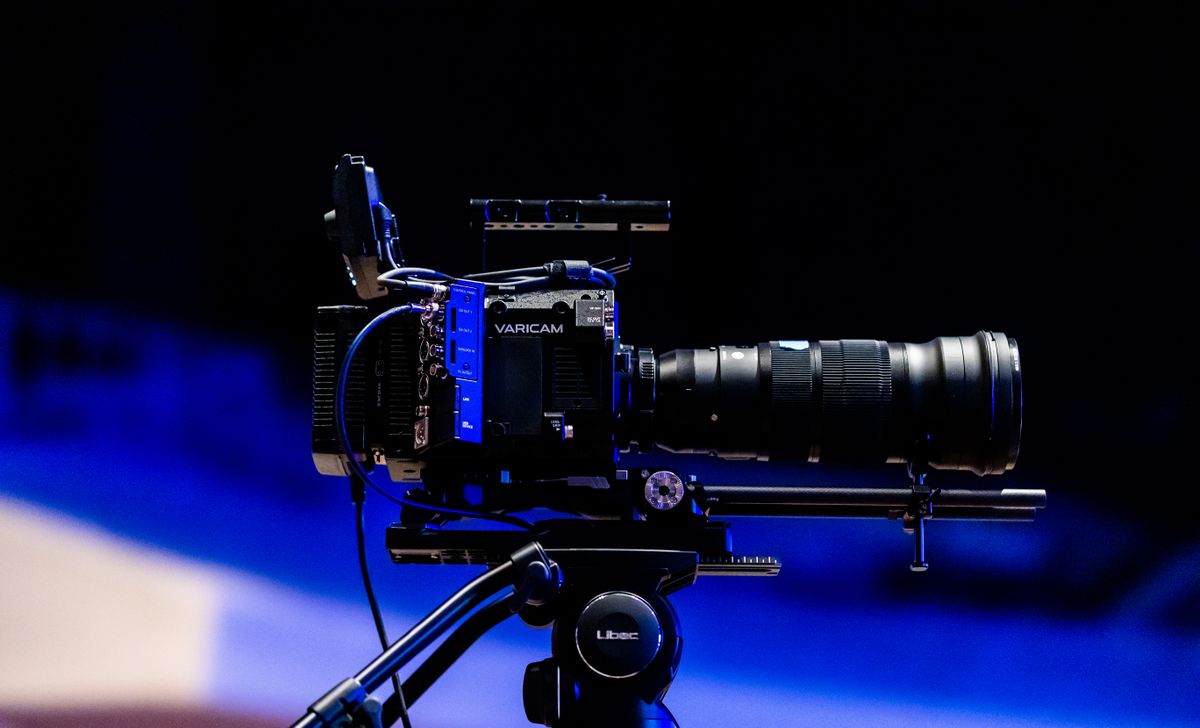 Kamera video profesional berlensa besar terpasang di tripod dengan latar biru gelap, siap untuk syuting