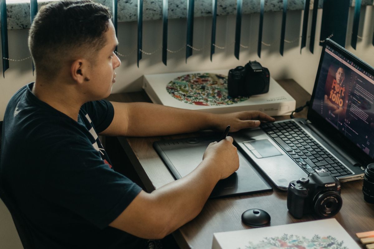 Seseorang mengedit desain di laptop memakai tablet gambar, menggambarkan proses mengedit poster di Canva