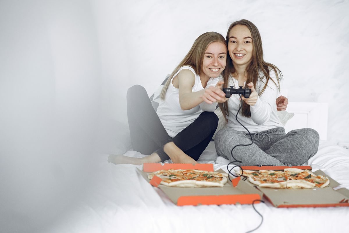 Dua perempuan duduk di tempat tidur sambil memegang stik game bersama dan tersenyum, dengan dua kotak pizza terbuka di depan mereka