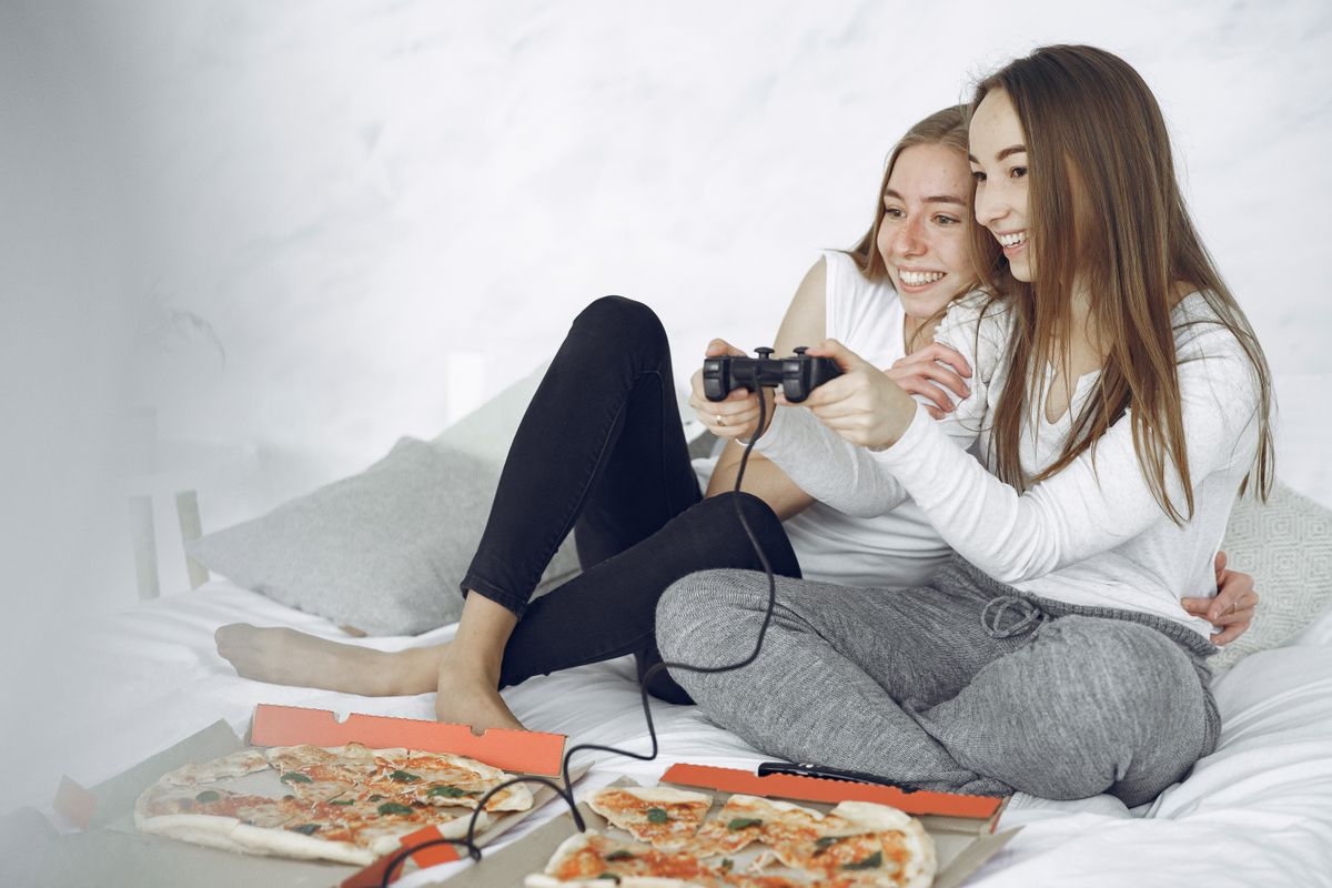 Dua perempuan tertawa sambil bermain game bersama dengan stik game di tangan dan kotak pizza terbuka di depan mereka