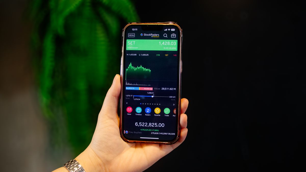 Tangan memegang HP yang menampilkan aplikasi trading saham dengan grafik harga real-time, menggambarkan jual beli saham yang kini bisa dilakukan dari ponsel