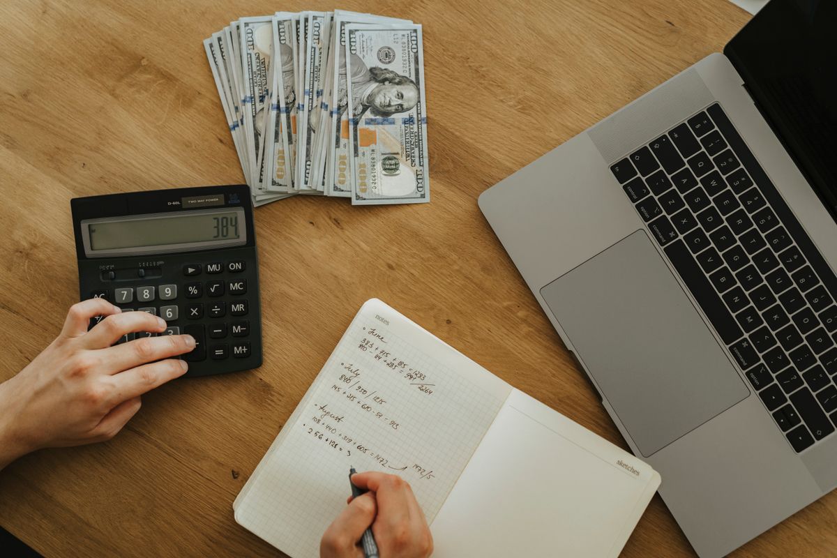 Tangan menekan kalkulator dan menulis rencana di buku catatan di samping uang dan laptop, menggambarkan persiapan anggaran sebelum mulai berinvestasi