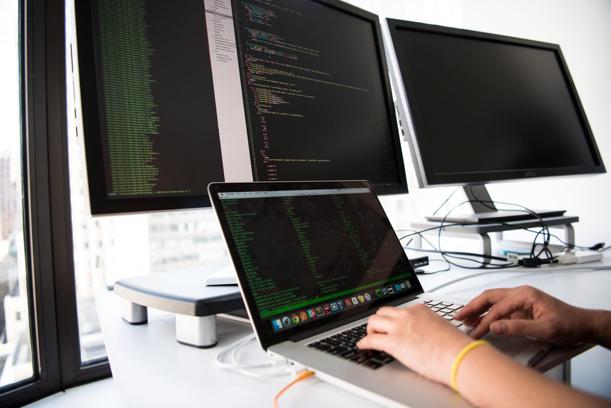Tangan mengetik di laptop yang layarnya menampilkan kode, dengan dua monitor tambahan berisi kode di belakangnya
