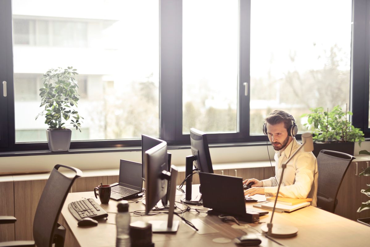 Seorang pria memakai headset sambil bekerja di depan beberapa komputer dan laptop di meja kantor dekat jendela