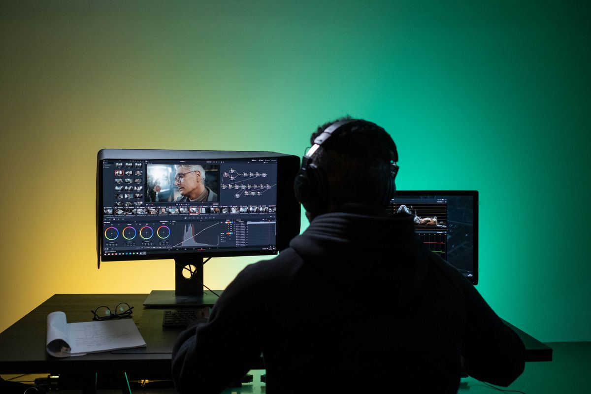 Seorang editor video memakai headphone sambil mengedit rekaman di dua monitor yang menampilkan linimasa dan roda warna