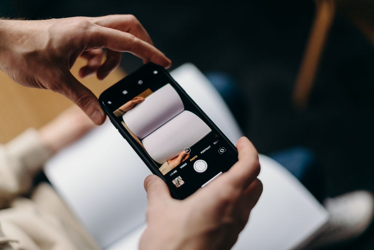 Dua tangan memegang HP yang kameranya diarahkan ke buku terbuka, menggambarkan proses memindai halaman dokumen di iPhone