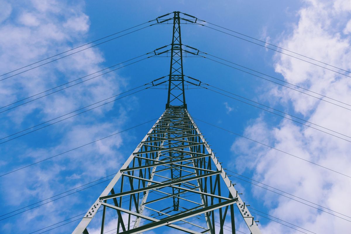 Menara transmisi listrik tinggi menjulang ke langit biru berawan, menggambarkan jaringan listrik PLN