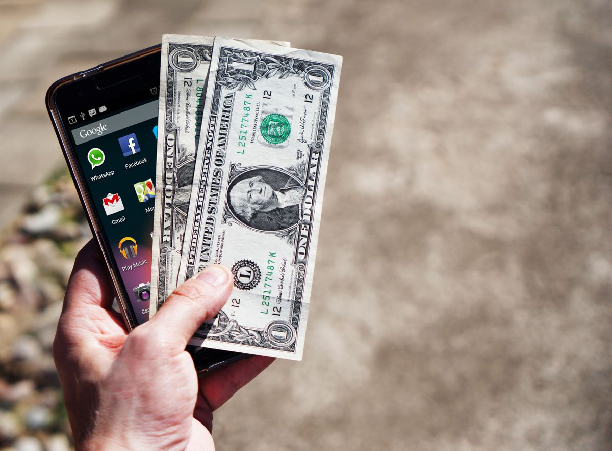 Tangan memegang HP dan dua lembar uang dolar Amerika Serikat, menggambarkan kirim uang ke luar negeri dari ponsel