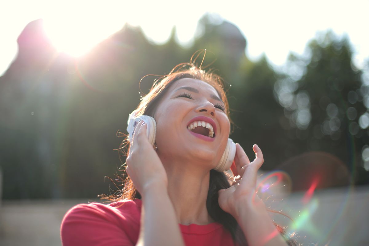 Perempuan berbaju merah tertawa sambil memakai headphone putih di luar ruangan dengan cahaya matahari