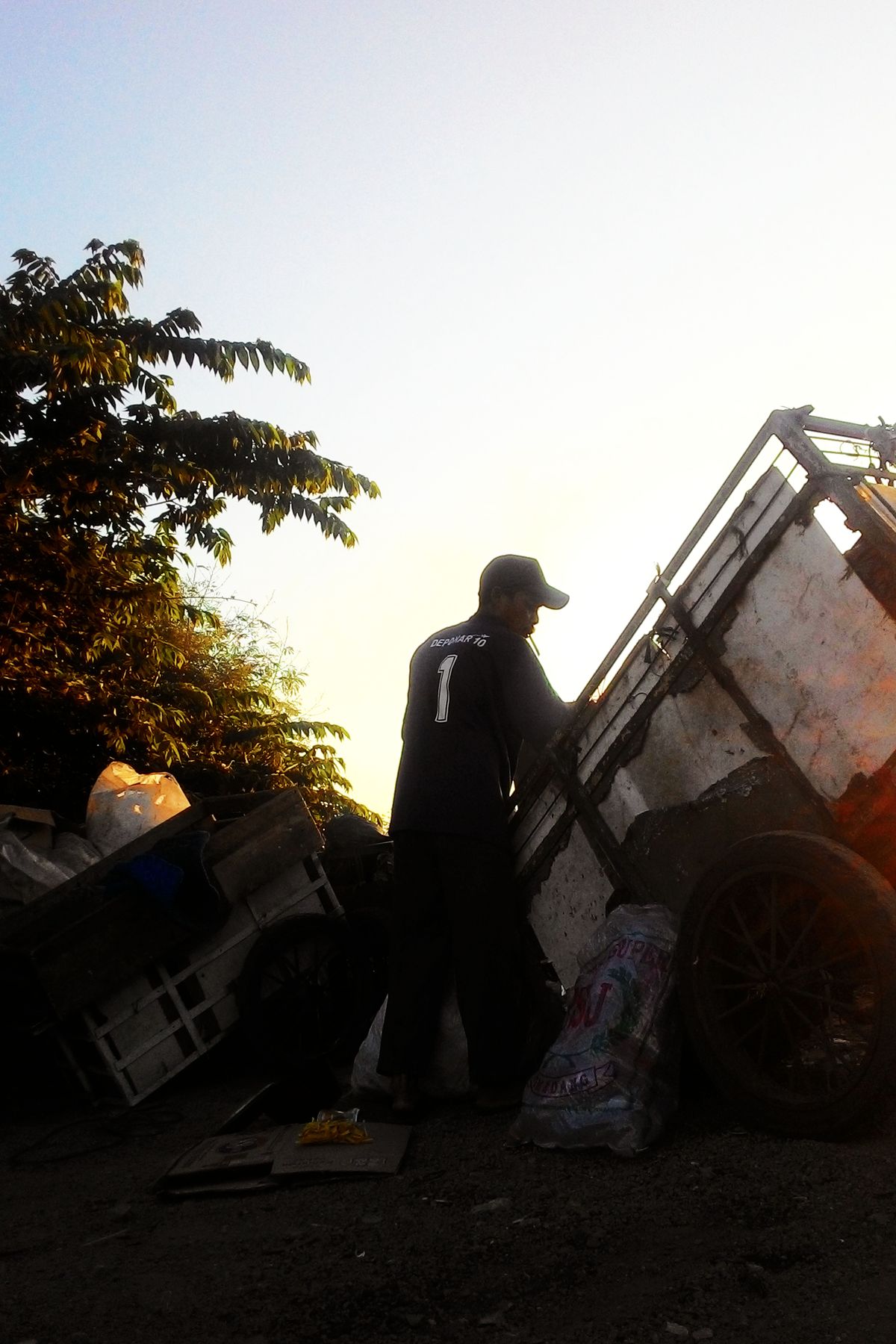 Siluet petugas kebersihan yang bekerja dengan gerobak sampah saat matahari terbenam di Bandung, menggambarkan pekerjaan lapangan penanganan sampah
