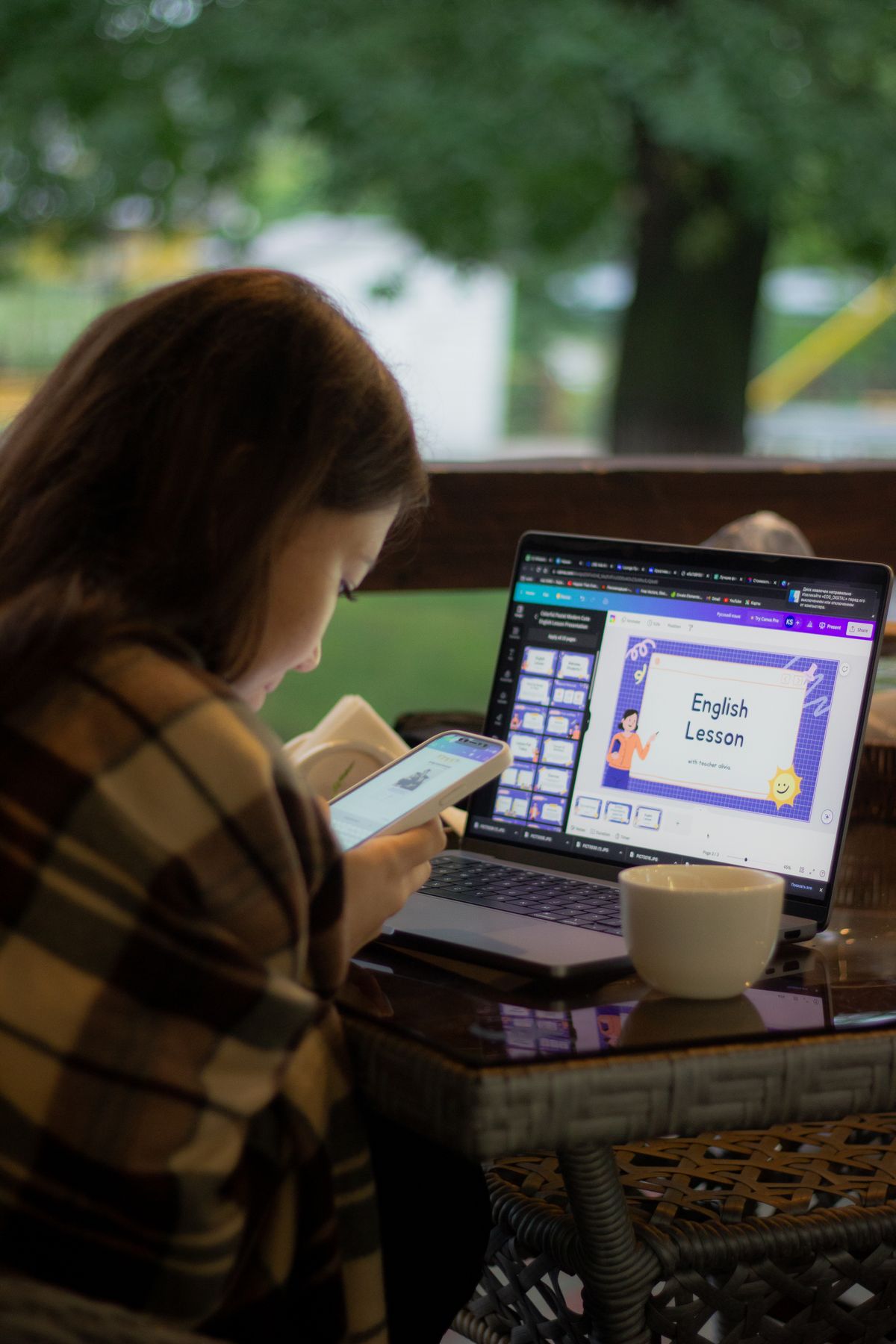 Perempuan mengikuti pelajaran bahasa Inggris di laptop sambil memegang HP, dengan secangkir kopi di meja