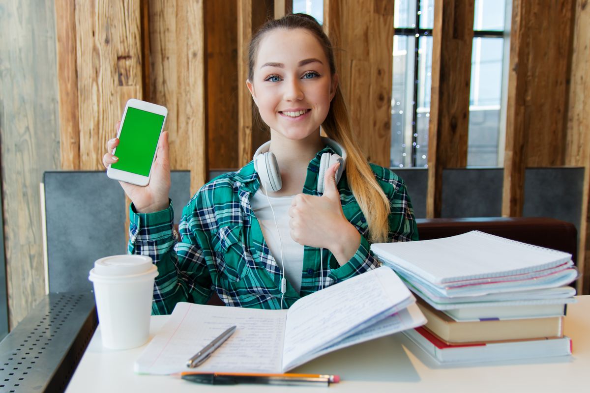 Perempuan tersenyum sambil memegang HP, dengan headphone di leher dan tumpukan buku di meja